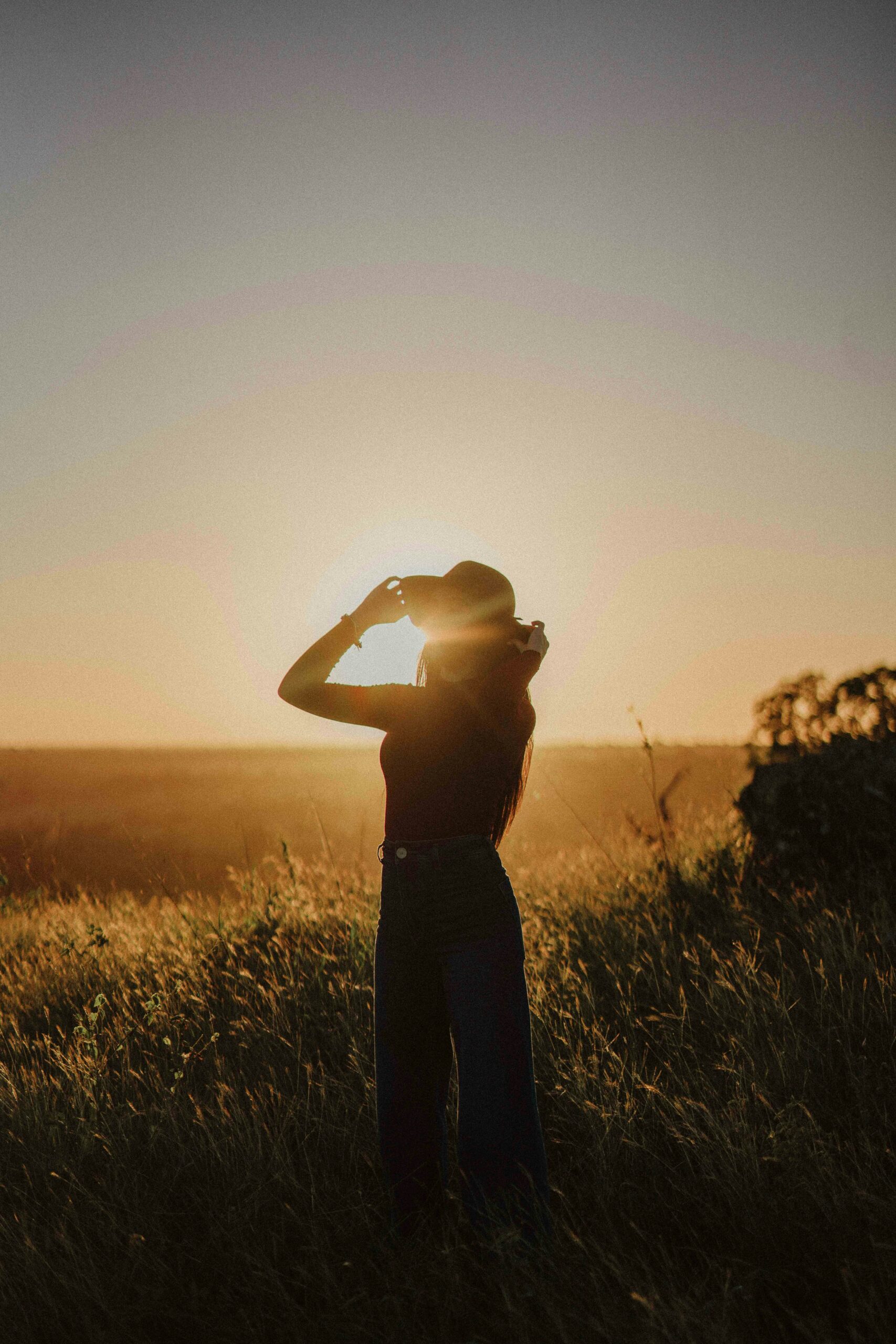 Silhouette of a woman standing in a field at sunset, lifting her hat as sunlight streams through, surrounded by tall grass and golden light.