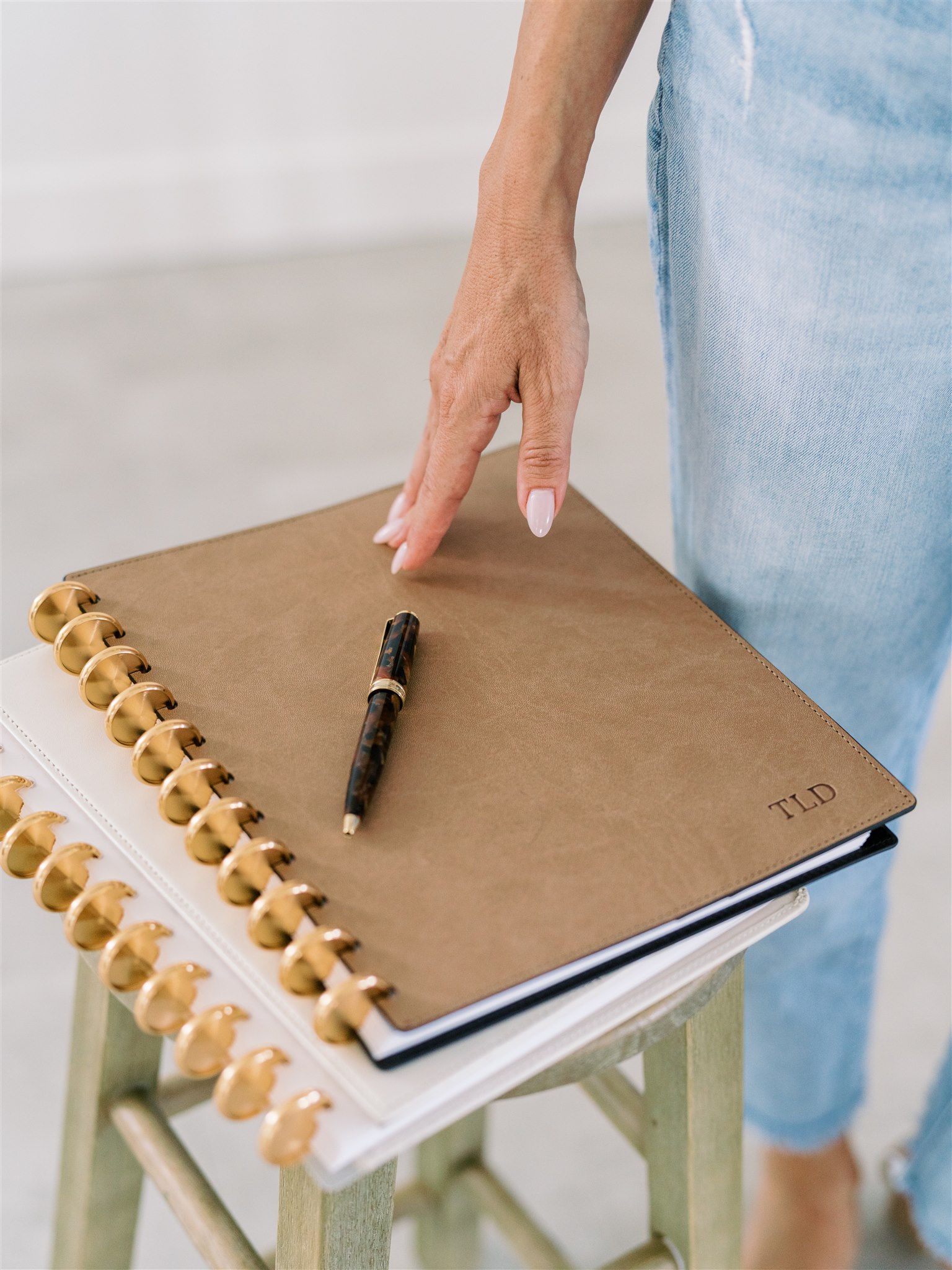 Woman’s hand resting on a personalized disc-bound journal with gold rings and pen, styled on a wooden stool with soft, natural light.