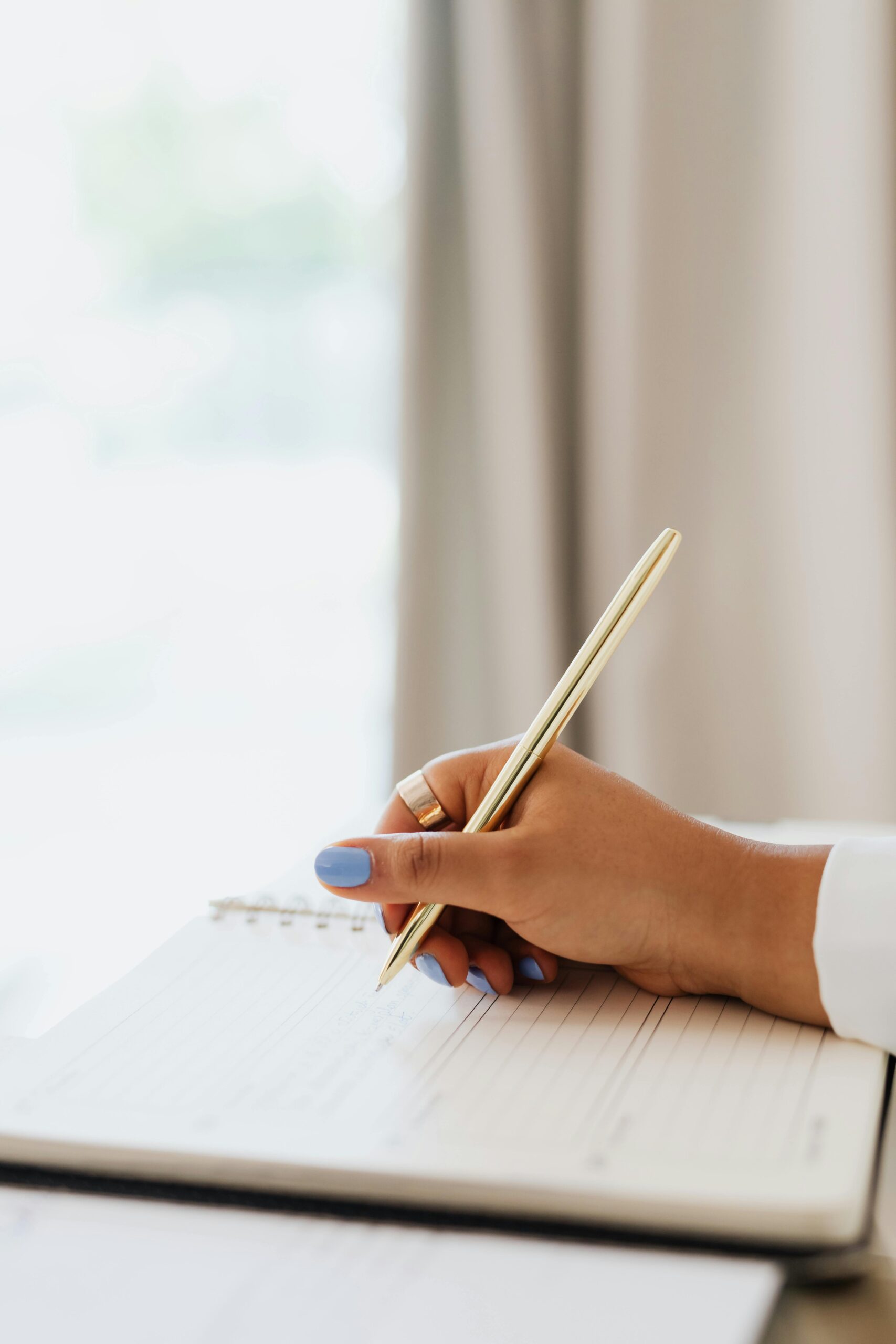 Close-up of a woman journaling with a gold pen on a lined notebook, her soft blue nail polish and rings reflecting a peaceful, intentional moment of reflection.